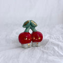 Ceramic sculpture of 2 tiny, cute cherry characters connected to the same stem. They have gold detailing.