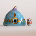 Decorative ceramic teardrop-shaped yurt sculpture with gold accents and a tiny character wearing a pink hood standing next to it.