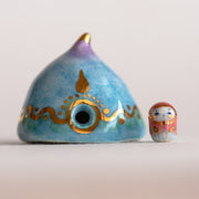 Decorative ceramic teardrop-shaped yurt sculpture with gold accents and a tiny character wearing a pink hood standing next to it.