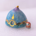 Decorative ceramic teardrop-shaped yurt sculpture with gold accents and a tiny character wearing a pink hood standing next to it.