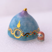 Decorative ceramic teardrop-shaped yurt sculpture with gold accents and a tiny character wearing a pink hood standing next to it.