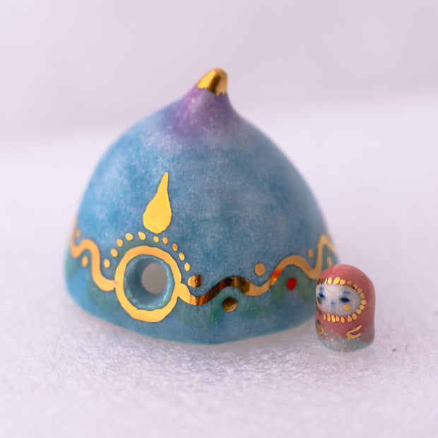 Decorative ceramic teardrop-shaped yurt sculpture with gold accents and a tiny character wearing a pink hood standing next to it.
