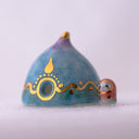 Decorative ceramic teardrop-shaped yurt sculpture with gold accents and a tiny character wearing a pink hood standing next to it.