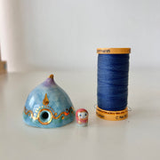 Decorative ceramic teardrop-shaped yurt sculpture with gold accents and a tiny character wearing a pink hood standing next to it. It sits next to a thimble for size reference.
