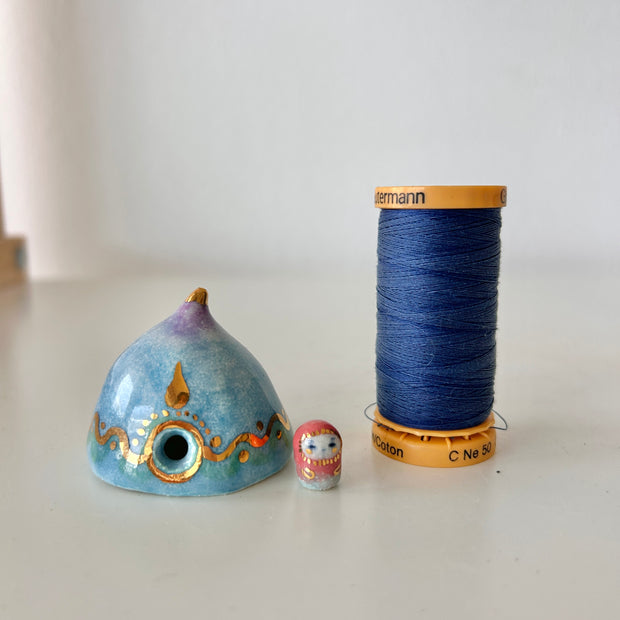 Decorative ceramic teardrop-shaped yurt sculpture with gold accents and a tiny character wearing a pink hood standing next to it. It sits next to a thimble for size reference.