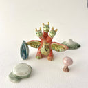 Small collection of sculptures: a three-headed monster, a small cherry blossom tree, green mountain and other nature elements.
