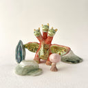 Small collection of sculptures: a three-headed monster, a small cherry blossom tree, green mountain and other nature elements.
