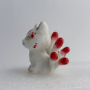 White ceramic cat figurine, sitting with 9 tails that are red-tipped. The cat's face and body has gold painted detailings.