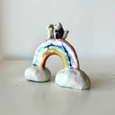 Small ceramic sculpture of a rainbow with clouds on the ends and 2 tiny foxes standing at the top, facing each other as though they are getting married.