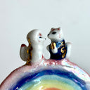 Small ceramic sculpture of a rainbow with clouds on the ends and 2 tiny foxes standing at the top, facing each other as though they are getting married.