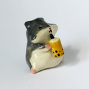 Ceramic sculpture of a gray and white hamster, sitting and holding a cup of thai tea boba.