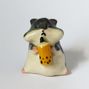 Ceramic sculpture of a gray and white hamster, sitting and holding a cup of thai tea boba.