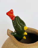 Unglazed brown ceramic pot with a green lizard character breaking out of it, with a tiny flame coming out of its mouth.