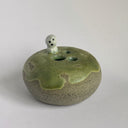 A handmade ceramic pot with a rounded, speckled gray-brown base and a glossy, dripping sage green glaze on top. A small, cute ghost figure with black eyes and a mouth sits on the rim.