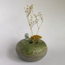 A handmade ceramic bud vase with a speckled gray base, a green glazed top, and a small white ghost figurine. It holds dried yellow and white flowers, set against a plain light gray background.