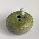 A handmade ceramic pot with a rounded, speckled gray-brown base and a glossy, dripping sage green glaze on top. A small, cute ghost figure with black eyes and a mouth sits on the rim.