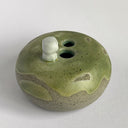 A handmade ceramic pot with a rounded, speckled gray-brown base and a glossy, dripping sage green glaze on top. A small, cute ghost figure with black eyes and a mouth sits on the rim.