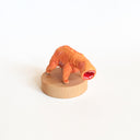 Small orange creature figure with four legs and no face, only an open mouth with many teeth. It stands on a wooden base against a white background
