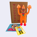 Orange vinyl figure with a bow tie and glasses, standing next to a cardboard box with Japanese text, on a light gray background.