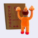 Orange toy figurine with red shoes in front of a cardboard box with Japanese text and illustrations.