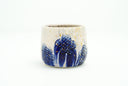Cream colored cup with blue flowers as a repeating design, they have grid patterning and their coloring bleeds down the side of the cup. Gold specks decorate the cup.