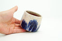 Cream colored cup with blue flowers as a repeating design, they have grid patterning and their coloring bleeds down the side of the cup. Gold specks decorate the cup.