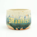 Ceramic cup with a glossy finish and an coloring of light yellow, teal blue and light orange that lightly bleeds into one another like a more abrupt, melty gradient. "Manifest" is written in a gold Old English font.