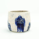 Cream colored cup with blue flowers as a repeating design, they have grid patterning and their coloring bleeds down the side of the cup. Gold specks decorate the cup.