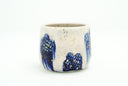Cream colored cup with blue flowers as a repeating design, they have grid patterning and their coloring bleeds down the side of the cup. Gold specks decorate the cup.