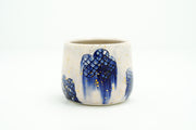 Cream colored cup with blue flowers as a repeating design, they have grid patterning and their coloring bleeds down the side of the cup. Gold specks decorate the cup.