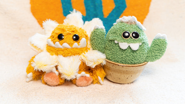 Plush sculpture of a cactus with large eyes and silly cartoon style overbite teeth. It has white claws on its hands and sits in a brown ceramic pot. It sits next to a plush cute monster with a flower collar around its head.