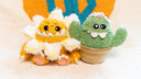 Plush sculpture of a cute, yellow monster with large cartoon style bead eyes and a smiling underbite. Around its head is a daisy flower made to look like a collar. It sits next to a plush green cactus with a smiling cartoon face.