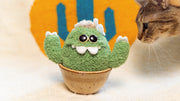 Plush sculpture of a cactus with large eyes and silly cartoon style overbite teeth. It has white claws on its hands and sits in a brown ceramic pot.