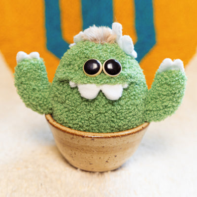 Plush sculpture of a cactus with large eyes and silly cartoon style overbite teeth. It has white claws on its hands and sits in a brown ceramic pot.