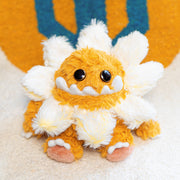 Plush sculpture of a cute, yellow monster with large cartoon style bead eyes and a smiling underbite. Around its head is a daisy flower made to look like a collar.