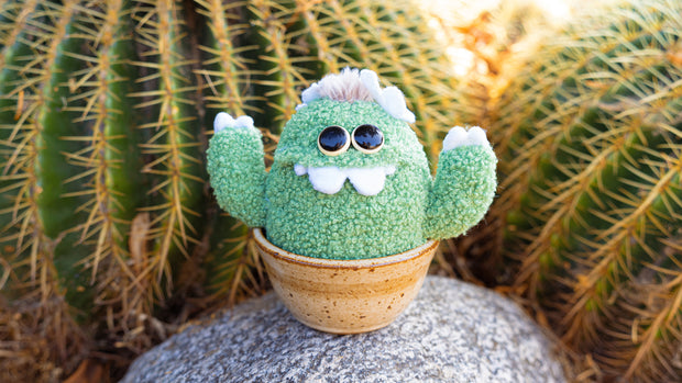 Plush sculpture of a cactus with large eyes and silly cartoon style overbite teeth. It has white claws on its hands and sits in a brown ceramic pot.