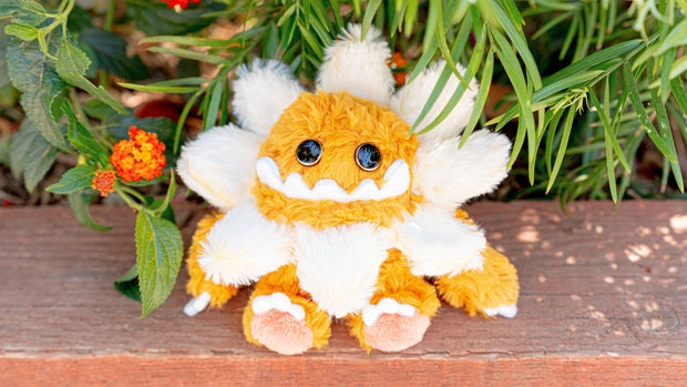 Plush sculpture of a cute, yellow monster with large cartoon style bead eyes and a smiling underbite. Around its head is a daisy flower made to look like a collar.