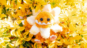 Plush sculpture of a cute, yellow monster with large cartoon style bead eyes and a smiling underbite. Around its head is a daisy flower made to look like a collar.