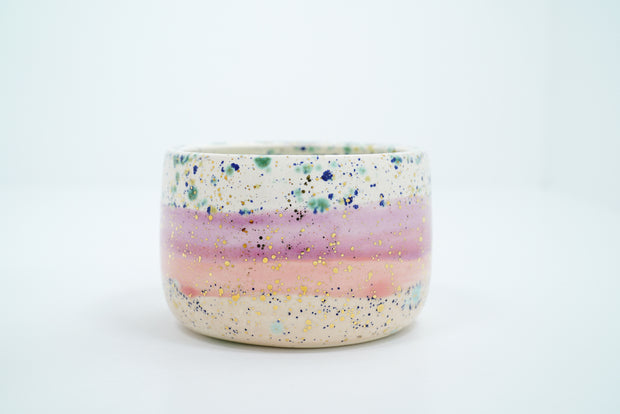 Short ceramic bowl, mostly off white with a pink stripe running through it and blue speckles all over. Gold specks are also on the surface.