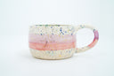 Short ceramic mug with thin large handle, mostly off white with a pink stripe running through it and blue speckles all over. Gold specks are also on the surface.