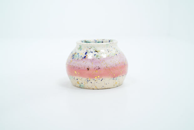 Small ceramic pot, with small opening mostly off white with a light pink stripe running through the middle. Blue and gold speckles decorate the surface.