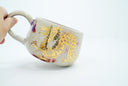 Ceramic cup with a thin handle, off white with subtle red splatter coloring. A gold dragon is painted onto the front of the mug.