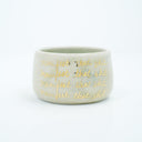 Short ceramic bowl, off white with four lines of repeated text that reads "manifest that shit" in gold cursive.