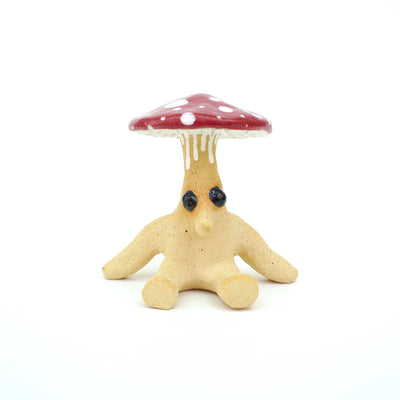Ceramic sculpture of a cartoon style mushroom, with a red and white cap and a brown anthropomorphic body and bulbous black eyes. It sits with its hands out to the side.