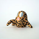 Small sculpture of a creature with large gray eyes and an 'o' shaped mouth, it is entirely brown with a repeating black squiggle pattern. Its face takes up most of its body and it leans back, on its 2 long arms and bends its long legs in front of it.