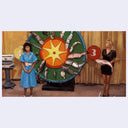 Two women standing on either side of a large wheel with fish, one holding a fish.