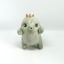 A small ceramic sculpture of a baby dinosaur with a textured light gray-green body, three gold horns on its head, and large yellow eyes, standing against a plain white background.