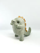 A small ceramic sculpture of a baby dinosaur with a textured light gray-green body, three gold horns on its head, and large yellow eyes, standing against a plain white background.