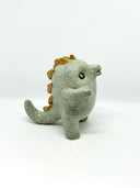 A small ceramic sculpture of a baby dinosaur with a textured light gray-green body, three gold horns on its head, and large yellow eyes, standing against a plain white background.