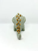 A small ceramic sculpture of a baby dinosaur with a textured light gray-green body, three gold horns on its head, and large yellow eyes, standing against a plain white background.